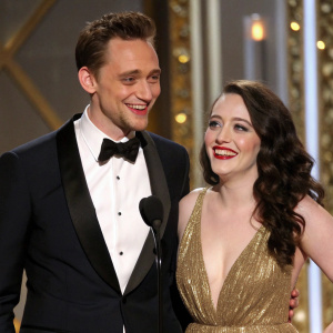 Tom Hiddleston and Kat Dennings attending a glamorous award ceremony. They both wear formal attire with matching warm smiles. Richly decorated backdrop with gold accents
