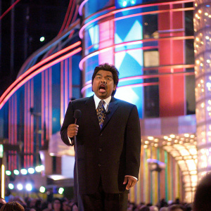 George Lopez at the 32nd Annual People's Choice Awards. Wearing a dark suit, white shirt, and patterned tie. Surprised expression on stage with colorful backdrop. Audience blurry in background.