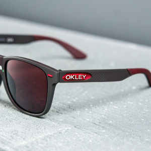 Close-up of Oakley sunglasses, dark matte frame, bold red OKLEY logo, curved arms, minimalist grey backdrop