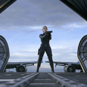 Katee Sackhoff as Kara 'Starbuck' Thrace. In a serious battle scene from Battlestar Galactica. She's in a black tactical outfit, looking determined. Background is a dark hangar with metal structures.