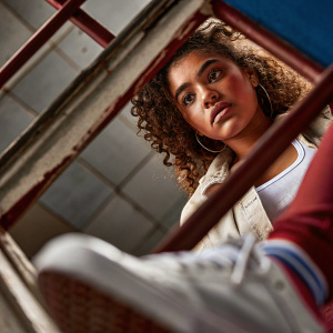 A high-resolution photo of a young Latina woman with curly hair. She is dressed in a casual outfit and has a serious expression. The blurred background has a cool blue tone, contrasting with her warm complexion. The focus is on her direct and engaging pose.