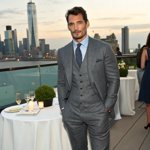 David Gandy, British model, dressed in tailored grey suit. Standing at posh fundraiser. City skyline in background.