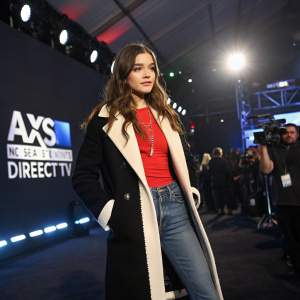 Hailee Steinfeld, young actress, standing at an indoor event with bright lights and camera flashes. She wears a black and white coat over a red top, jeans, and a necklace. The background is a dark stage with DirecTV and AXS TV logos.