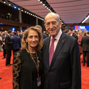 Ehud Olmert and Aliza Olmert, dressed formally, at a political conference. Crowded room with red carpeted floor.