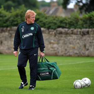 Gordon Strachan, wearing a Nike tracksuit with Carling logo, standing on a soccer field. He holds a soccer-themed duffel bag. Background includes a stone wall and green grass. Captured during a casual training session.
