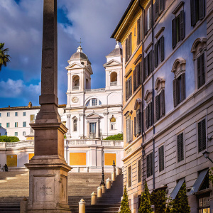 Photograph: Trinità dei Monti church, Rome, Italy. Tall stone pillar nearby. Big white building with intricate details. Blue sky with clouds.