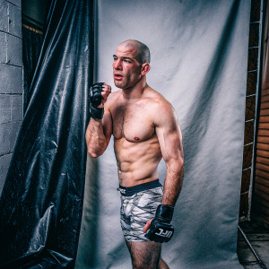 Dominick Cruz, UFC fighter, in a no-frills studio setting. Bald head, muscular frame. Wearing UFC shorts and gloves. Gray backdrop.