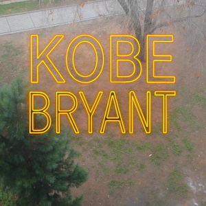 Dynamic graphic featuring 'KOBE BRYANT' in bold, stylized script. Neon yellow text against a deep violet backdrop.