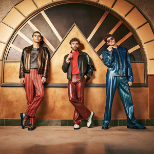 Years and Years band members Olly Alexander, Emre Turkmen, and Mike Goldsworthy, at the Brit Awards. Dressed in leather jackets and colorful attire. Q Awards logo and Absolute Radio branding backdrop.
