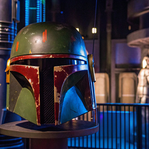 Boba Fett's Mandalorian helmet with green and red markings, displayed in a sci-fi exhibit.