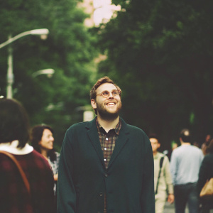 Jonah Hill, with stubble and friendly smile, wearing navy blue blazer over plaid shirt. Crowded street background