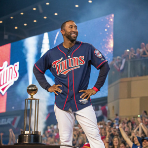 Eduardo Nunez, star player for the Minnesota Twins, poses confidently on the award stage. The backdrop is filled with cheering fans, creating a vibrant atmosphere. He wears his team's blue and white jersey, with the Twins logo prominently displayed.