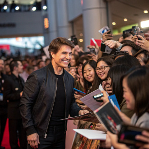 Tom Cruise, center stage at a crowded fan meet-and-greet event. Diverse crowd eagerly awaits autographs. Energetic atmosphere with fans of all ages, genders, and ethnicities.