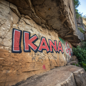High-contrast photo of the word 'Ikana' in graffiti style. The text is on a rugged rock face, close-up shot, showing sedimentary layers and rough texture.