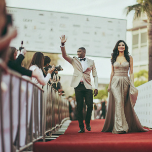 Larry English and Nicole Williams attending the Hollywood Film Awards. They stand confidently on the red carpet surrounded by a backdrop filled with various sponsor logos.