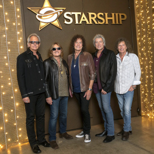 John Roth, Stephanie Calvert, Mickey Thomas, Darrell Verdusco, Phil Bennett, and Jeff Adams, members of Starship, at a gala event. They are standing in front of a gold backdrop with band logo. Party lights in background.