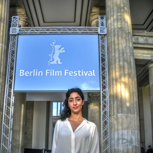 Golshifteh Farahani at the Berlin Film Festival, standing in front of a bright blue backdrop adorned with the official logo. She sports a classic white blouse and exudes a calm, composed demeanor.