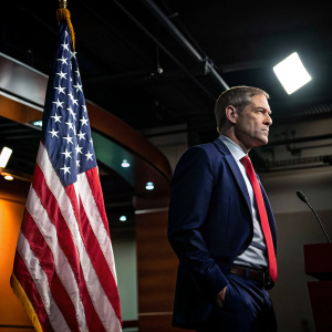 Jim Jordan, in a navy suit and red tie, stands beside the US flag at a press conference. Dimly lit room, spotlight on his face.