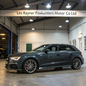Dark gray Audi A3 in showroom. Polished exterior under bright lights. 'Les Rayner Foxhunters Motor Co Ltd' backdrop with website info.