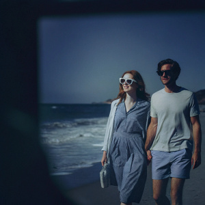Emma Stone and Andrew Garfield, dressed casually, walking on a sunny beach. Large sunglasses, relaxed poses