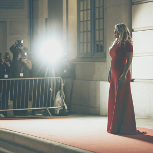 Kate Winslet at the BAFTA Film Awards. Wearing a long red dress. The background is a blur of flashing cameras and audience.