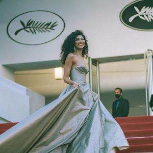 Zendaya, draped in a shimmering gold gown, stands confidently on the red carpet of the Cannes Film Festival. Her radiant smile and flowing, dark curls are the epitome of elegance against the stark, neon-lit backdrop featuring the festival's iconic palm tree logo.