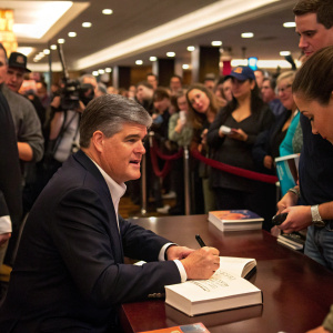 Sean Hannity, in a dark suit, signing autographs at a book signing event. Crowded room with many fans.