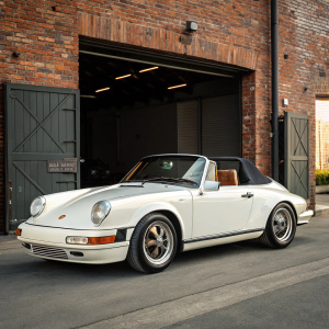 A pristine classic white Porsche 911 Carrera 2 Cabriolet from the late 90s, with its iconic rounded body and sleek design. Positioned in front of a brick wall and an open garage door. Under natural daylight, the car's details are clearly visible.