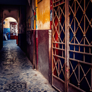 A narrow alleyway in a bustling medina. The ancient stone walls, lined with vibrant graffiti and faded signs. A rusty metal gate stands partially open, revealing a cobblestone path that leads deeper into the labyrinthine streets of Medina Sidonia, Cadiz Province, Andalucia, Spain.