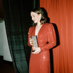Tessa Farmiga at the Gotham Awards. Wearing a sequined red dress, standing against a black backdrop. Holding a trophy