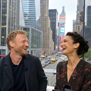 Kevin McKidd and Indira Varma laughing at a bustling press conference. Modern cityscape backdrop