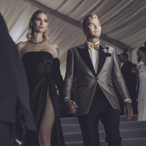 Chiara Ferragni and Fedez on the Met Gala red carpet. Chiara wears an elegant black gown with a high slit and plunging neckline. Fedez is in a sharp tuxedo with a golden-toned lapel and bow-tie, displaying tattoos. They are surrounded by other gala attendees.