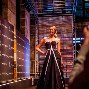 Candice Swanepoel, in a chic black strapless gown with a white accent line, posing confidently at the Mercedes-Benz Fashion Week. Textured backdrop with sponsor logos