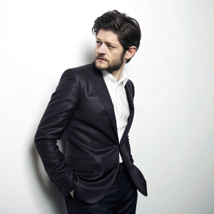 Iwan Rheon, ruggedly handsome with dark hair and a beard, stands confidently in a sleek black suit and white shirt. Shot against a plain white background. His stern expression captured in sharp focus.
