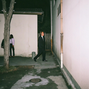 Michael Bolton, in a black suit, walking through a quiet, dimly lit alley at night. A casual passerby in the background. Simple, candid photo