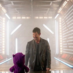 Taylor Lautner, looking jovial in a leather jacket, center stage at El Hormiguero. A purple plush toy in front. Futuristic, neon-lit backdrop.