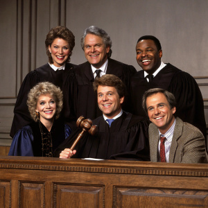 Reunion of 'Night Court' cast, Florence Halop, John Larroquette, Richard Moll, Charles Robinson, Markie Post, and Harry Anderson, posing for a promotional shoot. They are seated on a vintage wooden bench with a gavel in the middle, all dressed in period costumes.
