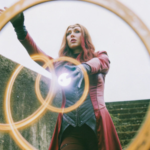 Wanda Maximoff, aka Scarlet Witch, stands in an action pose. She wields a glowing orb of energy. Her red, form-fitting costume clings to her figure. Long auburn hair flows behind her. Dark, void-like backdrop