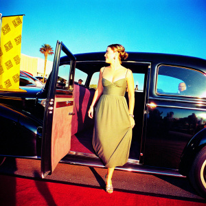 Kate Winslet steps out of a vintage black limousine at the star-studded gala. Wearing a sophisticated olive green dress with a fitted bodice and flowing skirt. Subtle silver jewelry complements her ensemble. She poses confidently on a deep red carpeted area with glitzy award show banners in the backdrop.