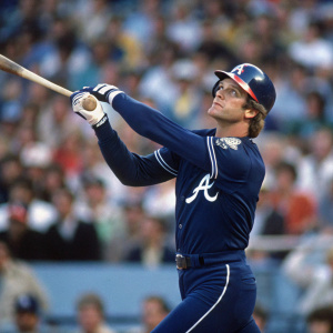 Dale Murphy in action, swinging a baseball bat at Dodger Stadium. Dark blue uniform with white 'A' logo. Crowd blurred in the background, highlighting the dramatic moment.