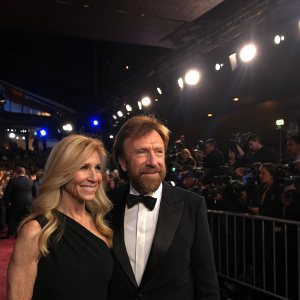 Chuck Norris and Diane Holechek attending an exclusive premiere event. They are standing in front of a crowd, with Norris dressed in a black tuxedo and bow tie. Dimly lit venue with flashing lights