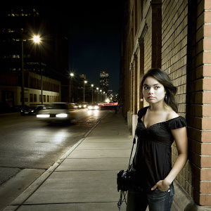 Vanessa Hudgens, looking intense, dressed in a black blouse and jeans. Stands in front of a busy city street at night.