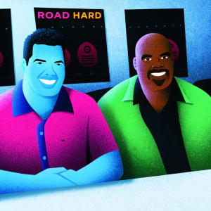 Adam Carolla and David Alan Grier at a promotional event. They are posing together with a friendly demeanor. The background is a plain white wall with 'Road Hard' movie posters and sponsor logos.