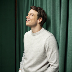 Jake Lacy, in a casual pose, wearing a light grey sweater. Studio backdrop featuring a green curtain.