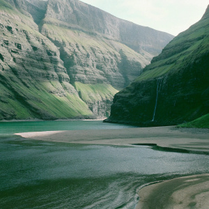 Stunning landscape of Saksun village in Faroe Islands. Majestic mountains loom over the tranquil grayish-sand beach. Dark green water contrasts with the rugged, layered rock formations of gray and green. A breathtaking photo.