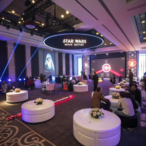 A lavish themed party for Star Wars fans. The room is filled with futuristic decor, including round tables and comfortable seating areas. The Star Wars Movie Battles logo is prominently displayed on the wall.