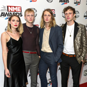 Ellie Rowsell, Joel Amey, Joff Oddie, and Theo Ellis from Wolf Alice pose seriously at the NME Awards. They are dressed in various outfits against a white backdrop with NME logos.