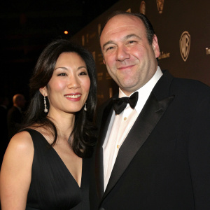 James Gandolfini and Deborah Lin at a gala event. Formal attire, black tuxedo, sleek black dress. They smile warmly at the camera. Dark background with subtle logos.