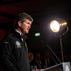 Rob Baxter, Exeter Chiefs Rugby Director, intense expression, curly hair, black jacket with logo. Formal event, dark room, spotlight on him