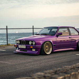 Custom-built BMW M3 E30 in vibrant purple paint with gold wheels. Digital illustration on asphalt, fence and ocean in blurred background.